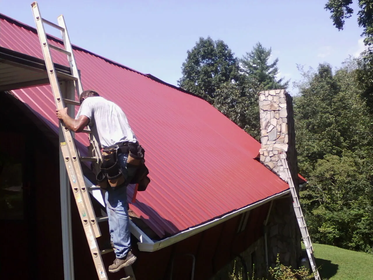 Roofer on a ladder beside a red metal roof and stone chimney — Soffit & Fascia Repair by Roofing Brothers in Shohola